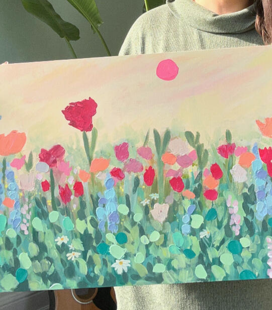 large canvas panel floral painting floral textured painting of a meadow in green, blue, pink, lilac and orange tones, in a semi abstract / modern impressionistic style. Pink sun in the sky. and a muted yellow background. painting is held by artist which is only partially visible in picture