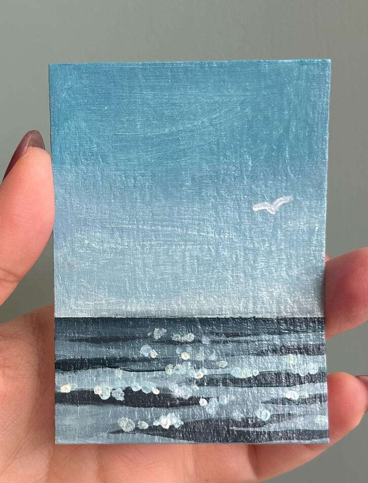 blue seascape aceo hand holding an aceo sized blue seascape painting on paper. featuring horizon, ripples, blue sky and a seagull silhouette in the sky